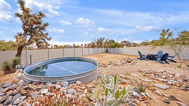 A swimming pool surrounded by rocks and desert vegetation, with a chair and table set up for outdoor dining. - Casa Cosmic: Hot Tub, Fire Pit & Stargazing (Joshua Tree)