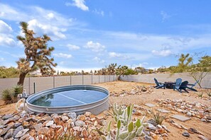 A swimming pool surrounded by rocks and desert vegetation, with a chair and table set up for outdoor dining.