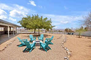 An outdoor seating area with turquoise Adirondack chairs arranged around a fire pit, a tree, and a view of the desert landscape.