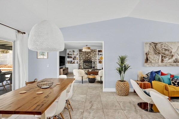 A modern living room with a wooden dining table, white chairs, a yellow sofa, and a large abstract painting on the wall.
