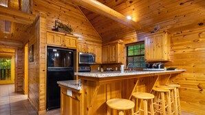 Une cuisine rustique aménagée dans une cabane en bois, avec un grand îlot central, des tabourets de bar, un réfrigérateur, un four à micro-ondes et un évier.