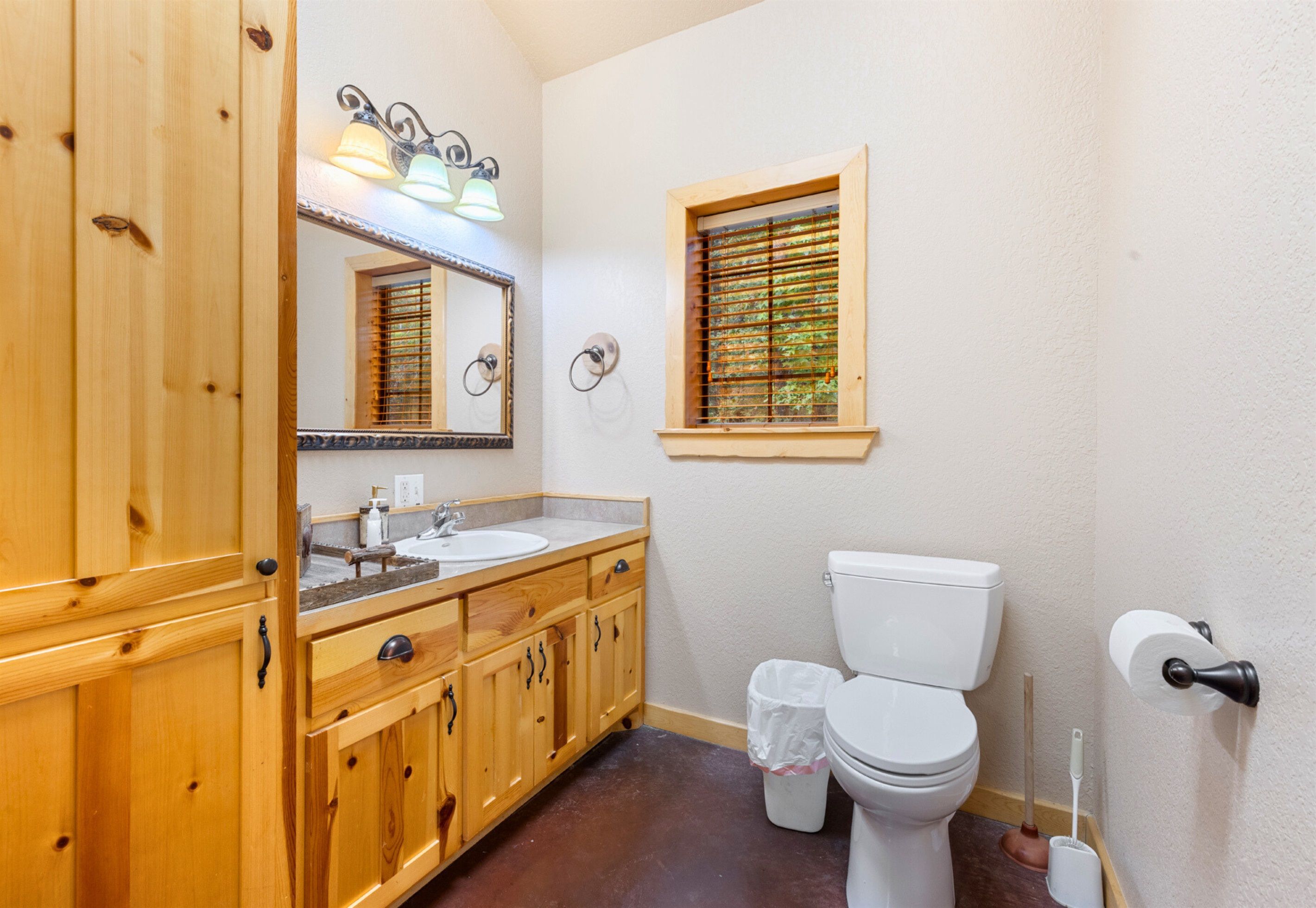 Une salle de bain avec des meubles en bois, un lavabo, des toilettes, une fenêtre avec des stores et un miroir avec éclairage.