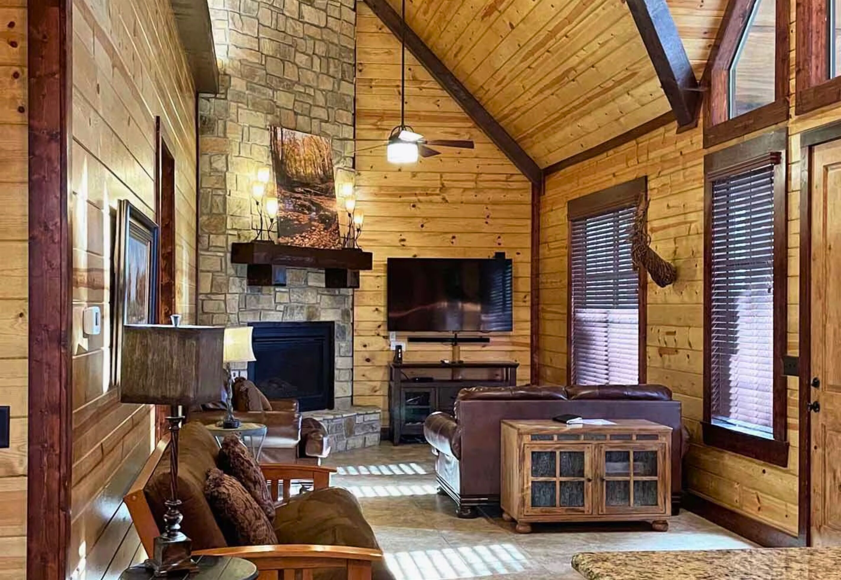 A cozy log cabin interior with a stone fireplace, leather sofa, and a television mounted on the wall.