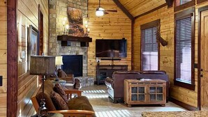 L’intérieur d’un chalet confortable, avec des murs en rondins de bois, une cheminée en pierre, un canapé en cuir et un téléviseur fixé au mur.