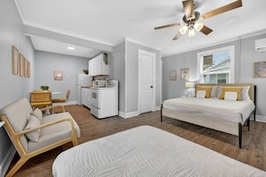 A bedroom with a bed, a chair, a desk, a kitchenette, and a ceiling fan.
