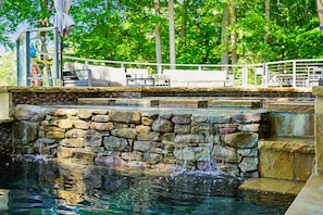 A stone waterfall pool with a stone wall, surrounded by outdoor seating and a white railing.