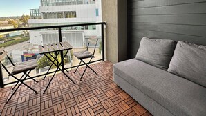 A balcony with a grey sofa, a wooden table, and a folding chair. The balcony has a herringbone-patterned floor and is furnished with hanging light bulbs. - Bright Modern Condo Near SkyTrain & Hospital (New Westminster)