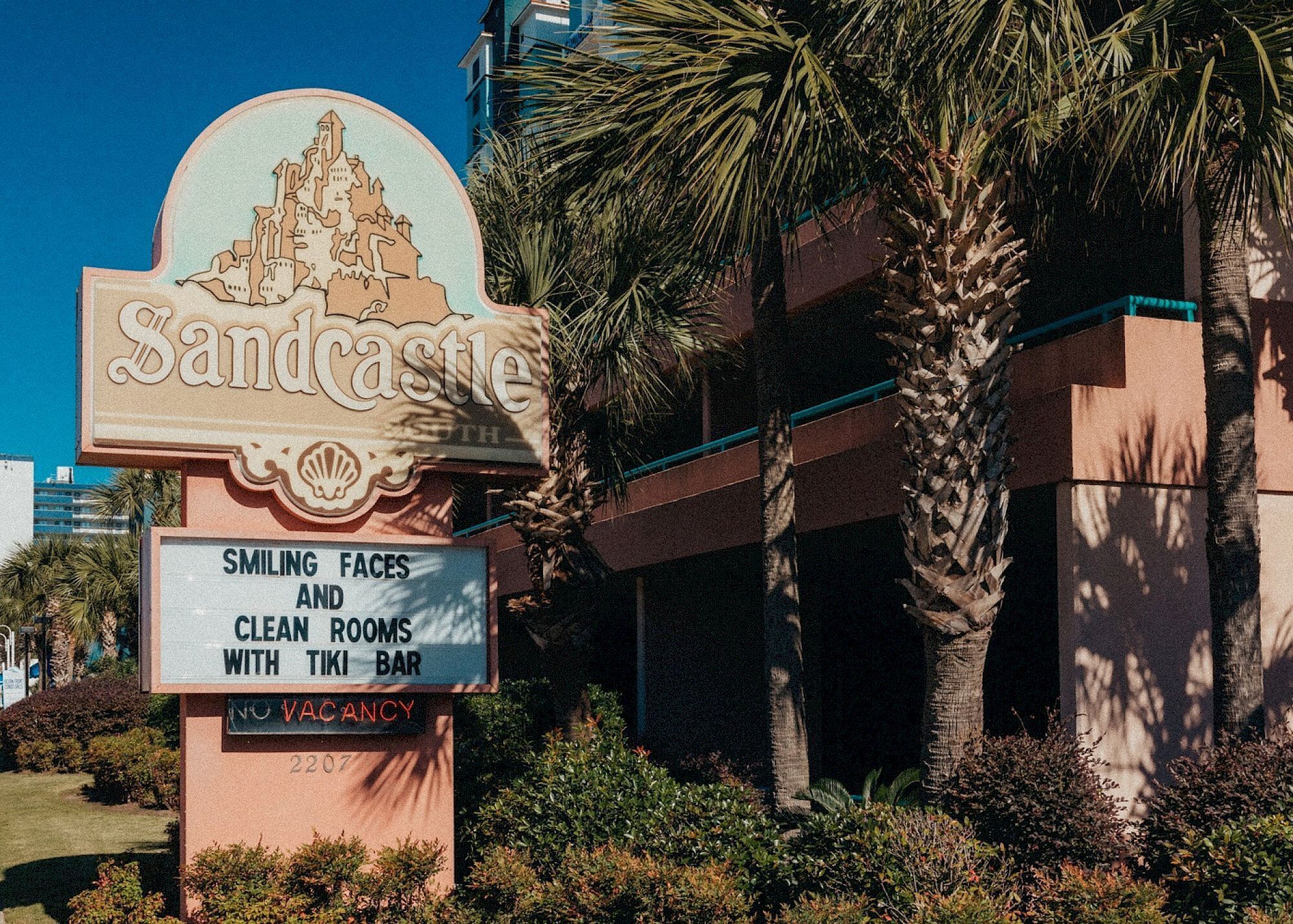 A hotel sign with a tiki bar, a no vacancy sign, and a palm tree in the foreground.