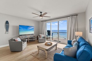 A living room with a TV, a ceiling fan, a sofa, a coffee table, and a view of the ocean through large windows.