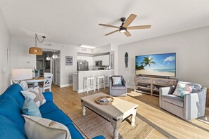 A modern living room with a blue sofa, grey armchair, and a wooden coffee table. A ceiling fan and a TV are visible.