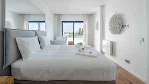 A modern hotel room with a large bed, a bedside table, a chair, a view of the city through large windows, and a decorative wall piece. - Stylish Algarve Retreat- Priv. Pool & Beach Nearby (Quarteira)