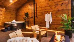 A wooden cabin interior with a sauna, a wooden bench, a fire pit, and a potted plant.