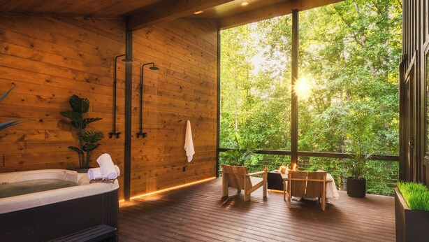 A wooden cabin with a hot tub, a dining area with chairs and a table, and a large window overlooking a forest.