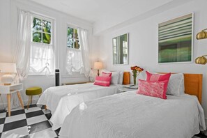 Quarto de hotel com duas camas, chão xadrez, escrivaninha com abajur e janela com cortinas leves.