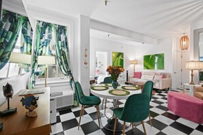 Uma sala de estar moderna com piso quadriculado, uma mesa de jantar redonda com cadeiras verdes, um sofá com almofadas coloridas e uma grande janela com cortinas tropicais.