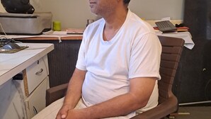A man sitting in a chair in a room with a desk, drawers, a keyboard, and a mug. - Narmada Guest House (Indore)