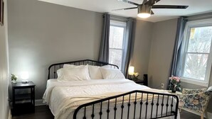 Ein ordentlich bezogenes Bett in einem Zimmer mit Deckenventilator, einem kleinen Nachttisch mit Lampe und einem stuhl mit Blumenmuster.