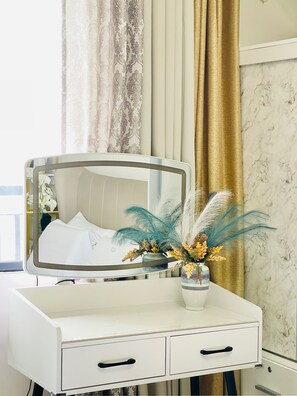 Un tocador blanco con espejo, un jarrón con flores secas y una cama con ropa de cama blanca al fondo.