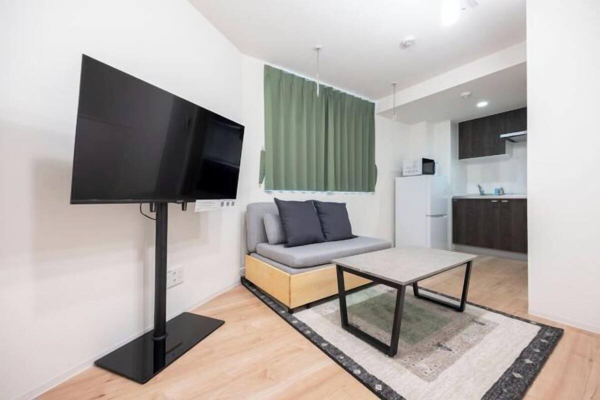 A modern living room with a flat-screen TV, a gray sofa, a small coffee table, and a kitchen area in the background.