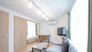 A modern living room with a sofa, a coffee table, and a TV mounted on the wall. There is a desk with chairs and a window with curtains. - MAT Nogata (Tokyo)