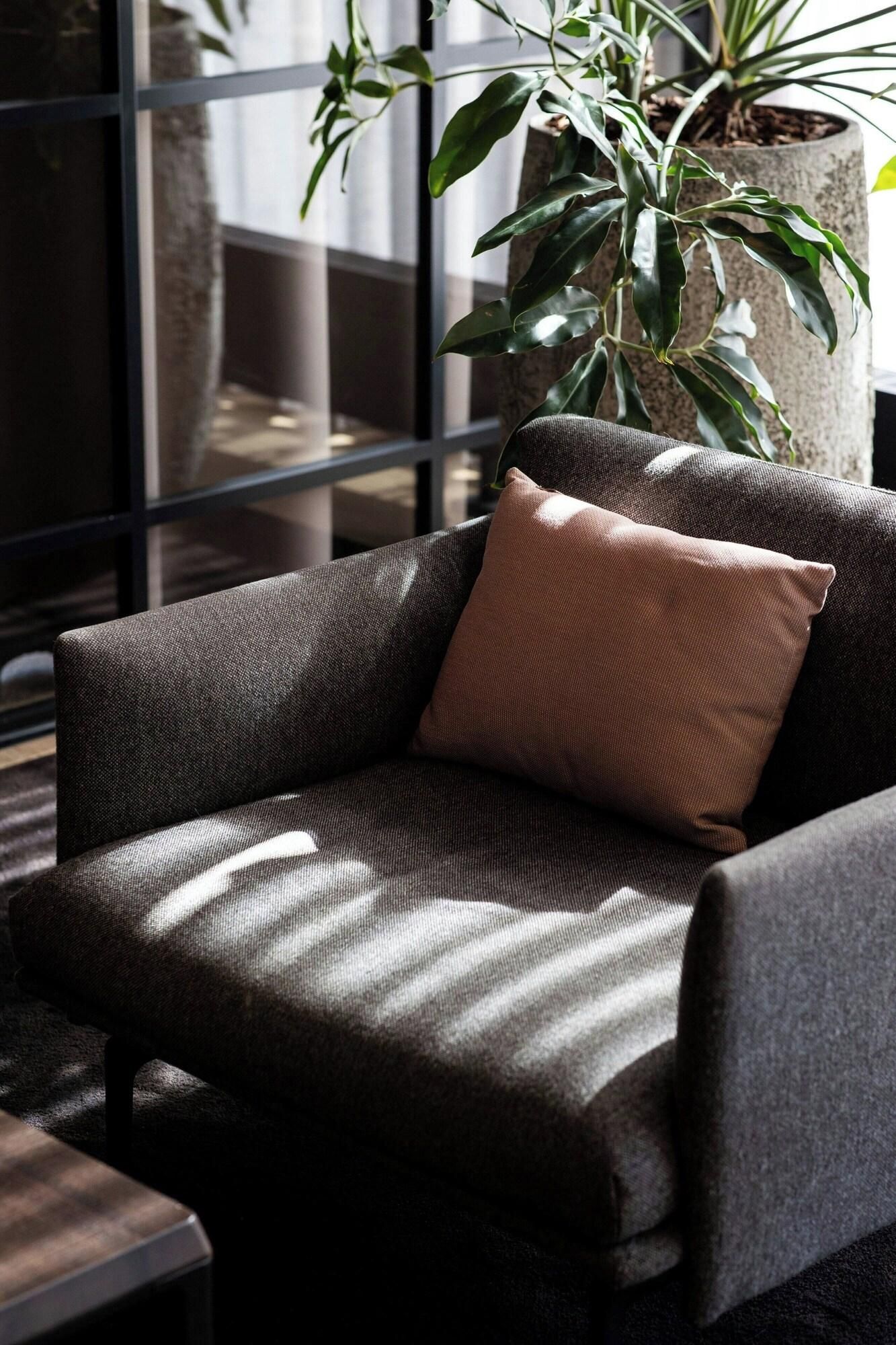 A modern armchair with a textured fabric and a single cushion, positioned in a room with a large window and a potted plant.