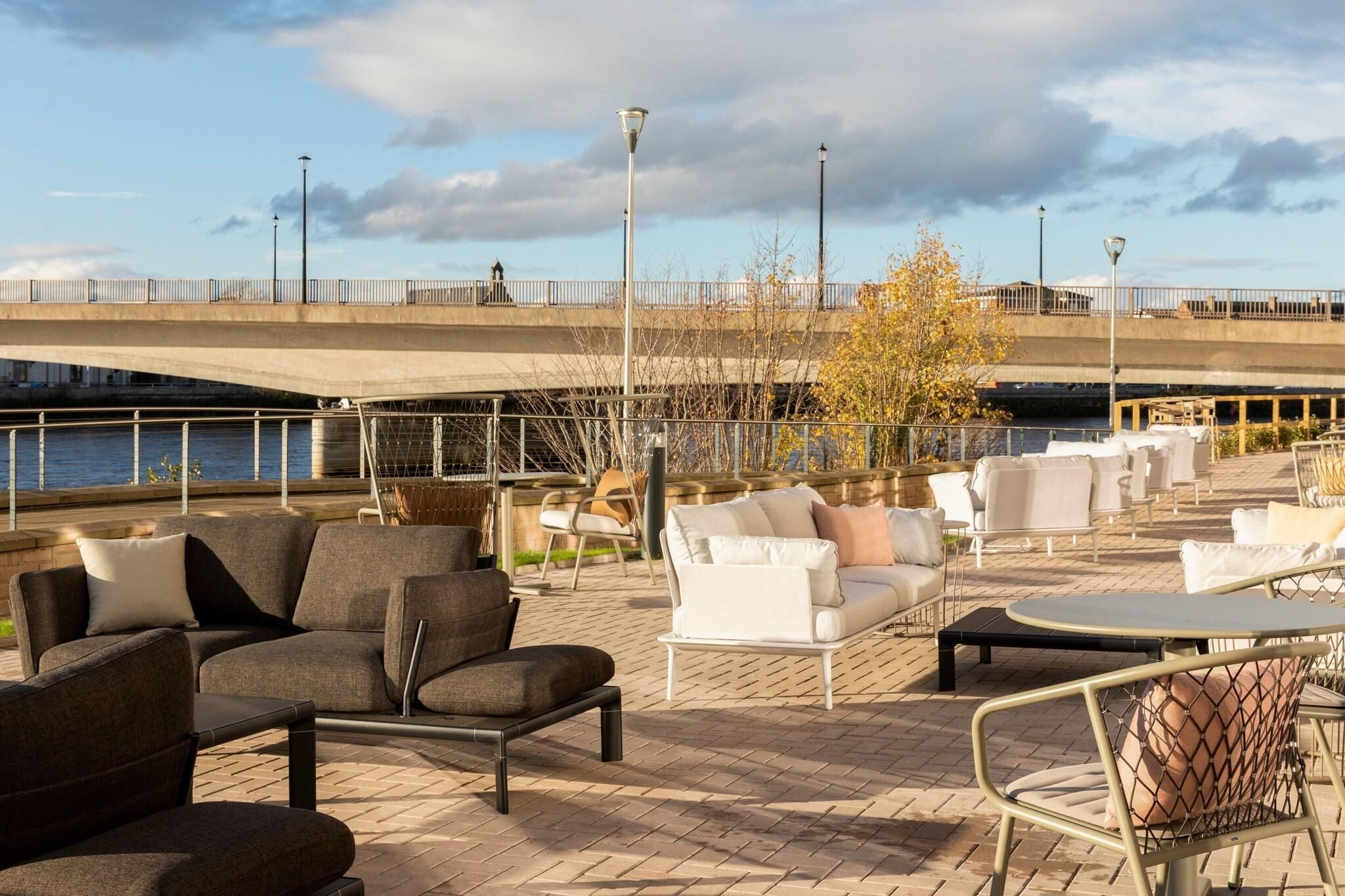 An outdoor seating area with various chairs and tables, a view of a river, and a bridge in the background.