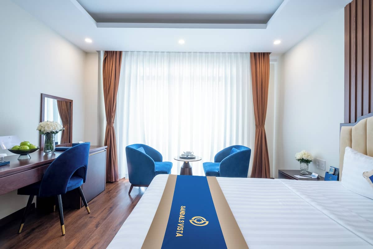 Habitación de hotel con cama, dos sillas azules, un escritorio con computadora y una ventana con cortinas.