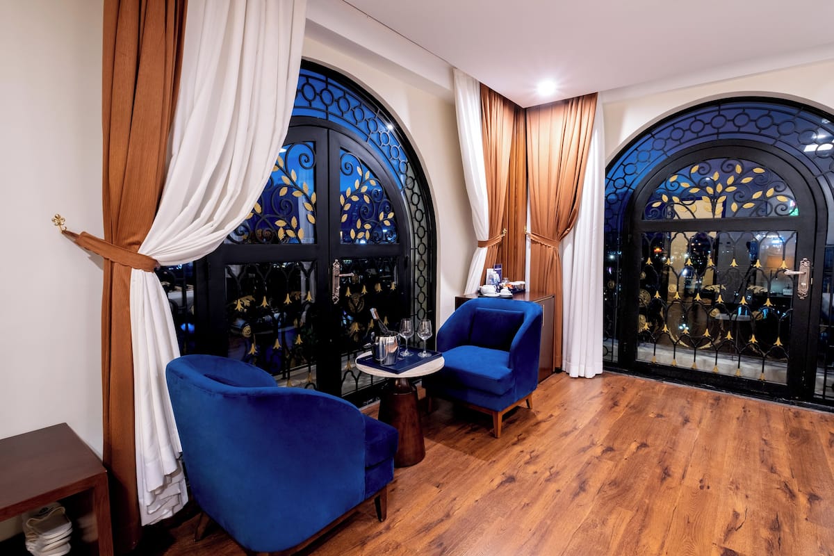 Una habitación con dos sillones azules, una mesita redonda pequeña y suelo de madera. Hay ventanas grandes con marcos negros decorativos y cortinas.