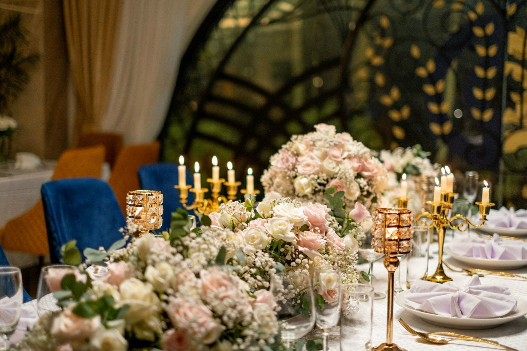 A table set with floral arrangements, candles, and gold candelabras in a room with blue chairs and a glass dome ceiling.
