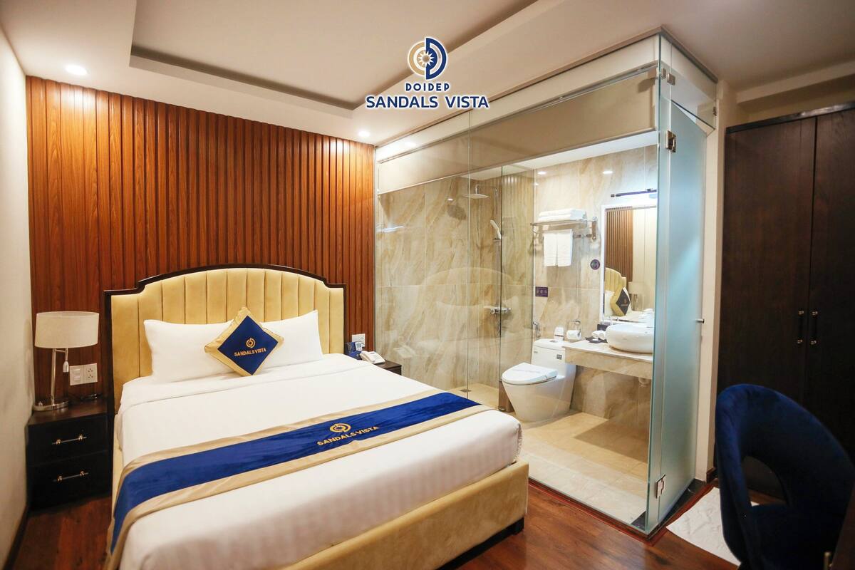 Habitación de hotel con cama, mesita de noche, ducha con paredes de cristal y un armario de madera oscura.