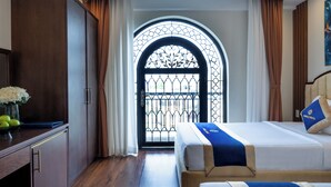 A hotel room with a large arched window, a bed with a blue and white bedspread, a nightstand with a vase of flowers, and a wooden wardrobe. - Sandals Vista (1 Bao Loc)
