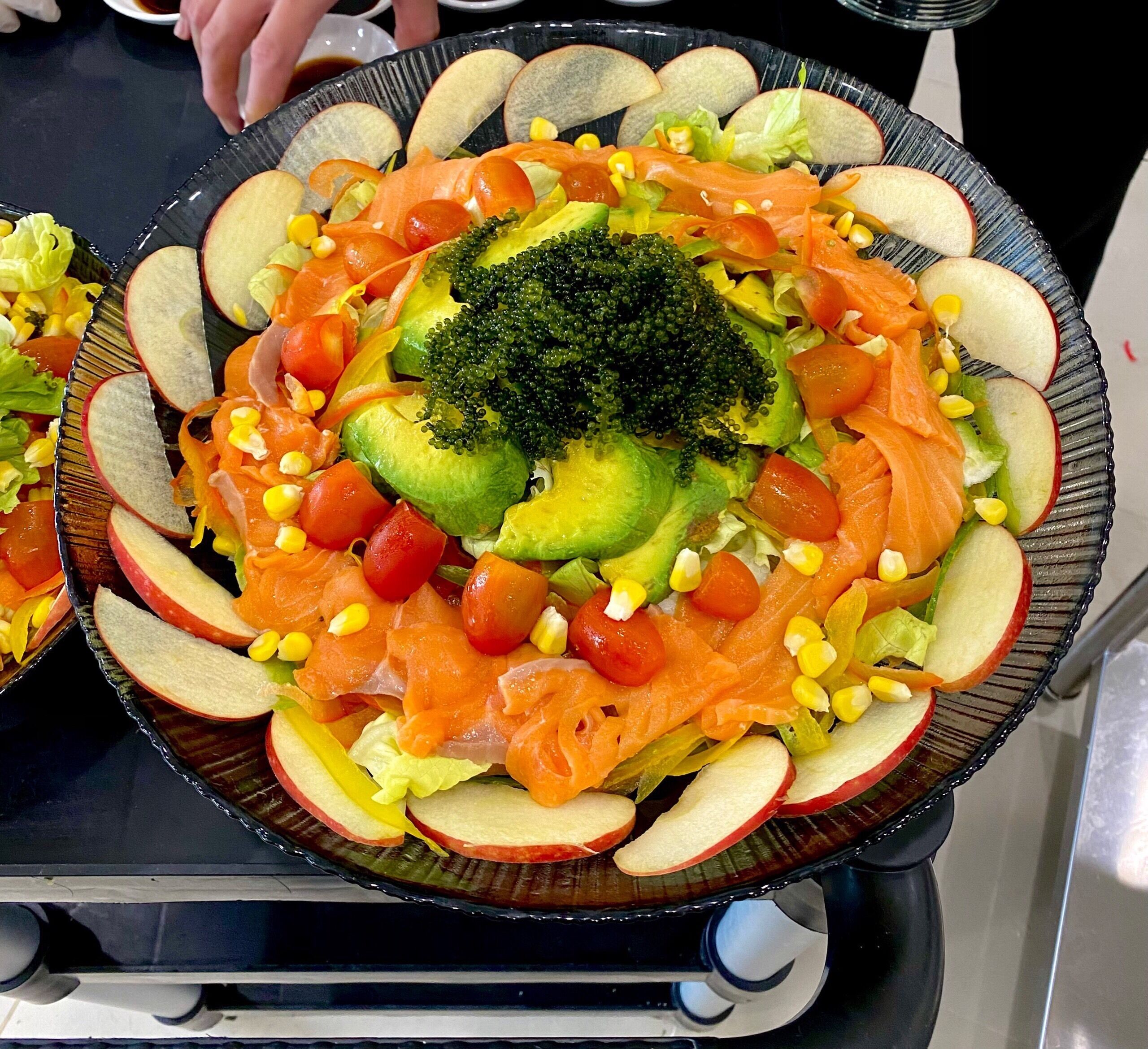 A large, round, dark-colored platter with a variety of sliced fruits and vegetables, including apples, tomatoes, and broccoli, arranged in a circular pattern.