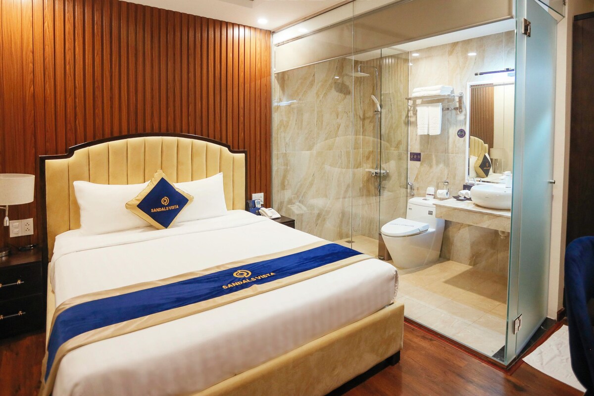 A hotel room with a bed, a nightstand, a glass-enclosed shower, and a dark wooden wardrobe.