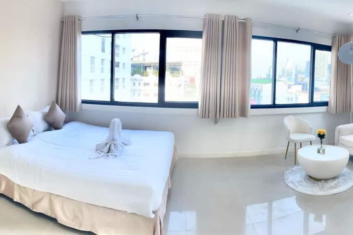 A modern hotel room with a large bed, a round coffee table, and a view of the city through large windows.