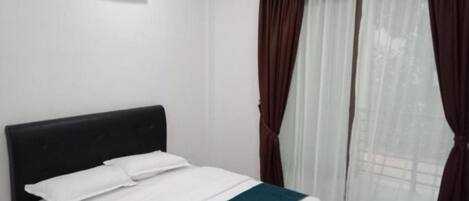 A single bed with a white mattress and pillows, a dark headboard, and a window with sheer white curtains and dark drapes.