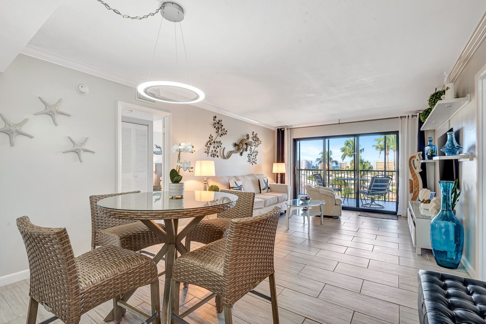 Ein modernes Wohnzimmer mit einem runden Glastisch, Korbstühlen, einer Couch und einem Balkon mit Blick auf Palmen.