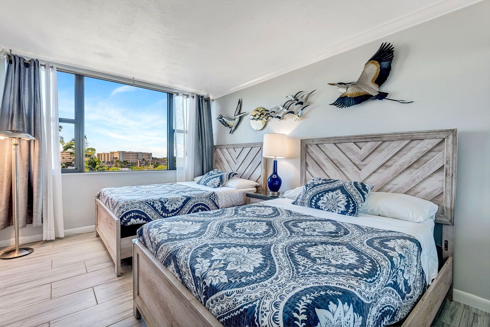 A hotel room with two beds, a large window with curtains, a lamp, and wall art including a bird and a skull.