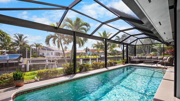 A swimming pool with a glass enclosure, a seating area with a sofa, and a view of a residential area with palm trees and houses. - 3BR Waterfront Home – Heated Saltwater Pool & Dock (Punta Gorda)