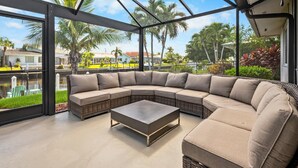 A modern outdoor living area with a curved sectional sofa and a coffee table, large glass windows, and a view of a residential area with palm trees and houses. - 3BR Waterfront Home – Heated Saltwater Pool & Dock (Punta Gorda)