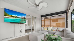 A modern living room with a large flat-screen TV, a ceiling fan, and a large window with blinds. - 3BR Waterfront Home – Heated Saltwater Pool & Dock (Punta Gorda)