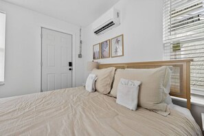 A bedroom with a bed, white walls, a door, and an air conditioning unit.