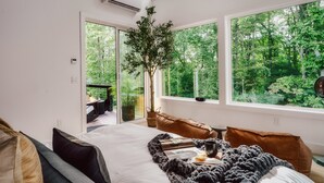 A bedroom with a large bed, a view of a forest through large windows, and a balcony with outdoor furniture.
