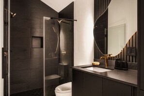 Une salle de bain moderne avec une douche à l’italienne en verre, des toilettes blanches et un meuble-lavabo aux tons foncés doté d’un robinet doré.