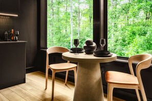 Un espace repas moderne avec une table ronde, deux chaises en bois et une vue sur une végétation luxuriante grâce à de grandes fenêtres.