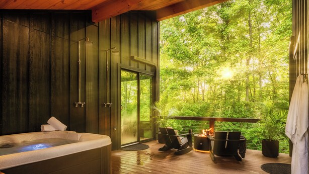 A hot tub in a wooden room with a glass door leading to a deck with outdoor seating and a fire pit.