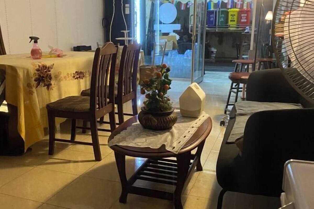 A dining area with a table covered by a yellow tablecloth, wooden chairs, a small potted plant, and a fan.