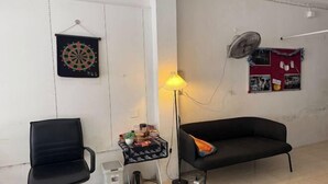 A room with a black office chair, a black sofa, a small table with a lamp, and a dartboard on the wall. - Sleep Break Hostel Sathon (Bangkok)