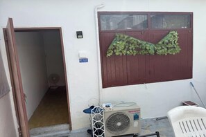 A room with a door leading to a hallway, a wall-mounted air conditioning unit, and a white plastic chair. - Sleep Break Hostel Sathon (Bangkok)