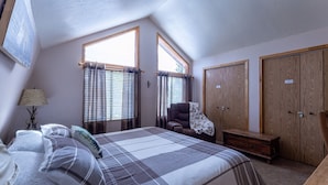Un dormitorio con una cama grande, dos ventanas con cortinas, un armario de madera y una silla.