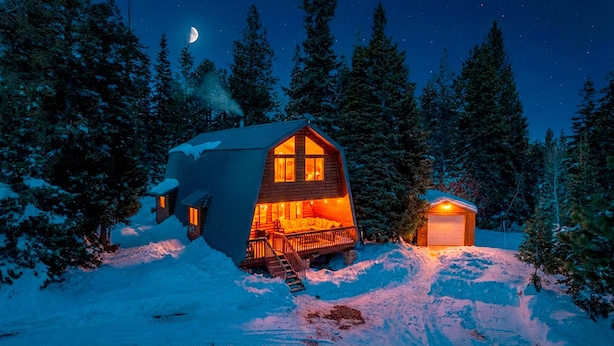 Una cabaña con una amplia terraza y un garaje, rodeada de nieve y árboles.